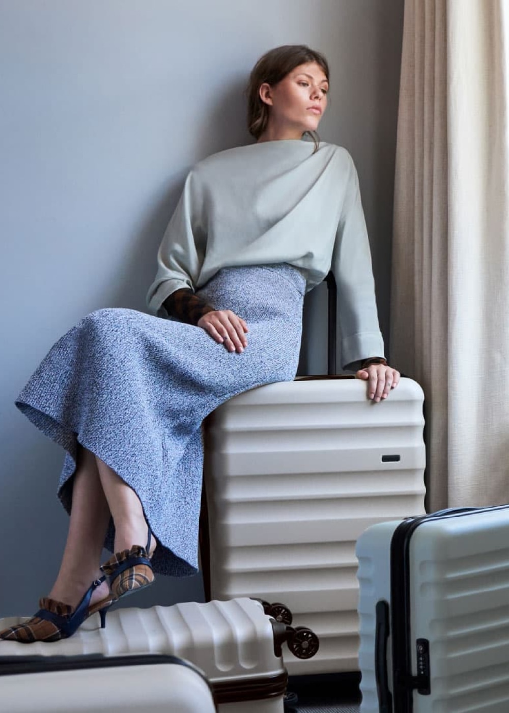 A woman sitting on a hard-case suitcase looking out a hotel window.