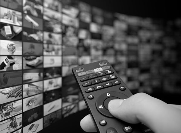 A person pointing a TV remote control at a wall screens, each displaying different TV channels and streaming content options.