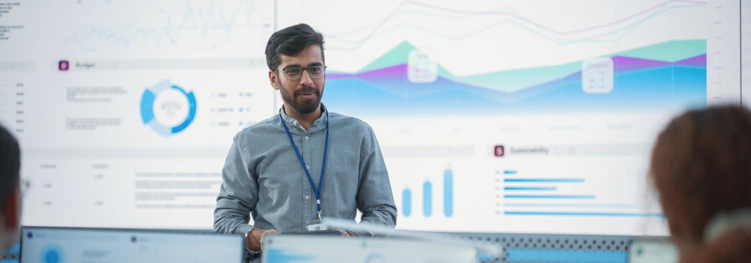Male Computer Scientist With Tablet Giving Presentation to Diverse Team Of Data Analysts In Front Of Big Digital Screen In Monitoring Room. Colleagues Listening To Training On Automation.