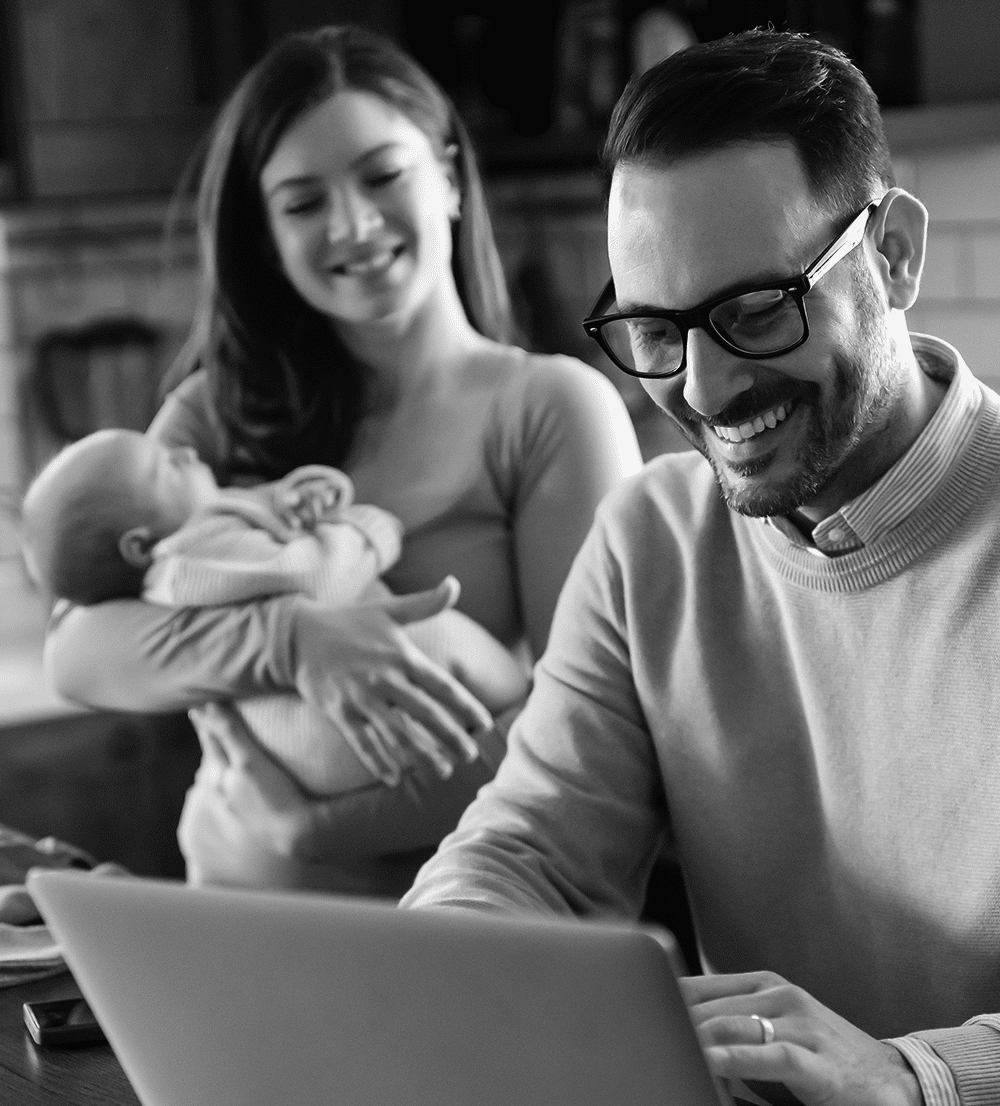 A man smiles as he looks at his superannuation balance on a laptop screen while a woman holding a baby stands behind him.